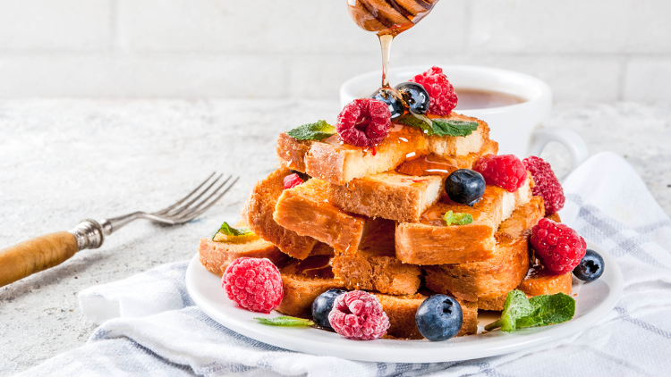 Ein Teller mit gestapeltem French Toast, garniert mit Beeren, Minzblättern und Honig, daneben eine Tasse und eine Gabel.