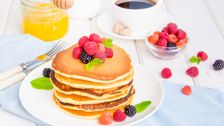 Ein Teller mit gestapelten Pancakes, garniert mit Himbeeren, Brombeeren und Minzblättern, daneben Honig, Kaffee und Früchteschale.