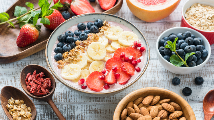 Eine Schale mit Haferflocken, Bananenscheiben, Heidelbeeren, Erdbeeren, Granatapfelkernen und Müsli, dekoriert mit Minze.