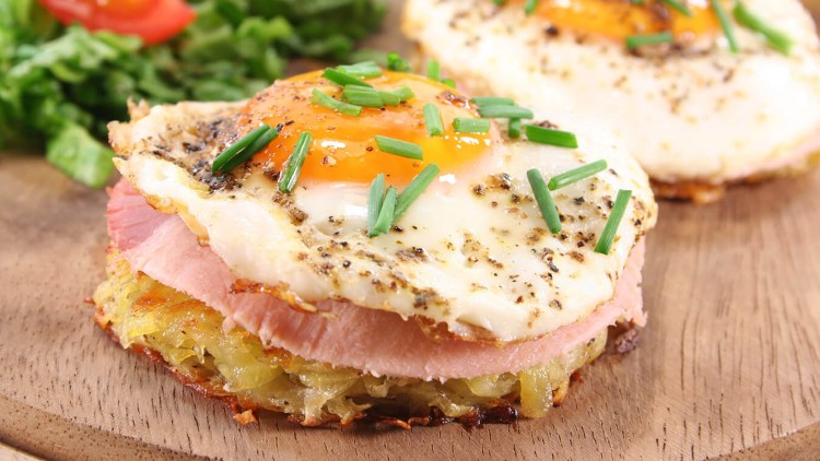 Ein Rösti-Taler belegt mit Schinken, Spiegelei, Schnittlauchringen und Pfeffer, im Hintergrund Salat und halbierte Tomaten.