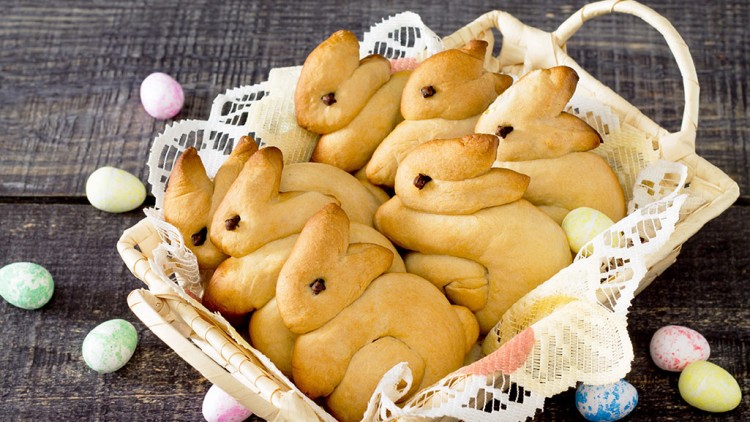Geflochtener Korb mit Hefegebäck in Form von Hasen mit Nelkenaugen auf hellem Stoff, daneben bunte, gesprenkelte Eier.