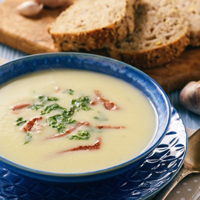 Cremige Suppe in einer blauen Schale mit Kräutern und Streifen, daneben ein Holzbrett mit geschnittenem Brot und Knoblauch.
