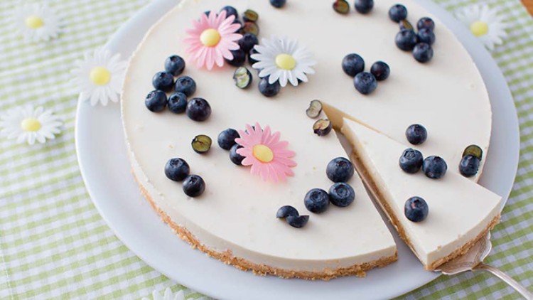 Runder Kuchen mit hellem Belag, dekoriert mit Blaubeeren und essbaren Blumen, ein Stück wird auf einem Tortenheber serviert.