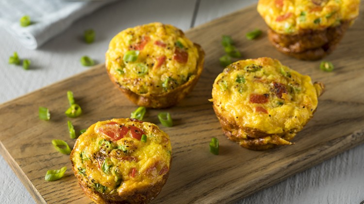 Vier goldbraune Eimuffins mit Tomaten, Brokkoli und Frühlingszwiebeln auf einem Holzbrett, dekoriert mit geschnittenen Zwiebeln.