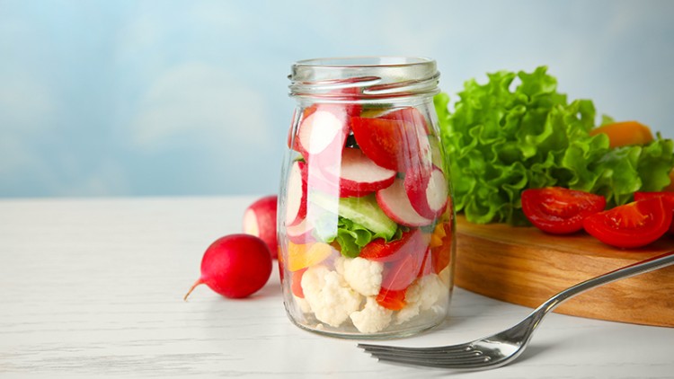 Ein Einmachglas gefüllt mit frischem Gemüse wie Radieschen, Salat und Blumenkohl steht auf einem Tisch, daneben liegt eine Gabel.