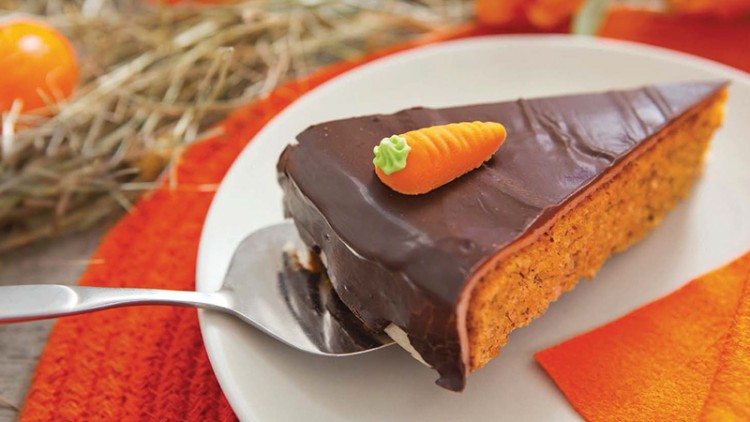 Ein Stück Schokokuchen mit Marzipan-Karotten-Deko auf weißem Teller, daneben Stroh, orange Serviette und kleine Kürbisse.