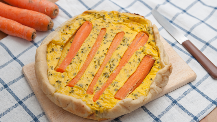 Eine herzhafte Tarte mit orangefarbenen Karottenstreifen auf gelber Masse liegt auf einem Holzbrett mit kariertem Tuch.