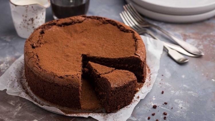 Runder Schokoladenkuchen mit Kakaopulver bestäubt, ein Stück herausgeschnitten, daneben Teller, Besteck und Kaffeegeschirr.
