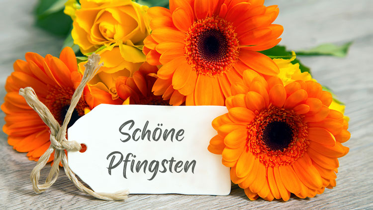 Ein Strauß aus orangefarbenen und gelben Blumen liegt auf hellem Holz mit einer Karte beschriftet mit „Schöne Pfingsten“.