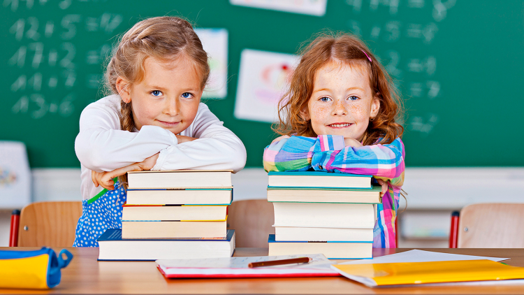 Zwei Stapel bunter Bücher auf einem Tisch vor einer grünen Tafel, mit Kinderzeichnungen im Hintergrund.