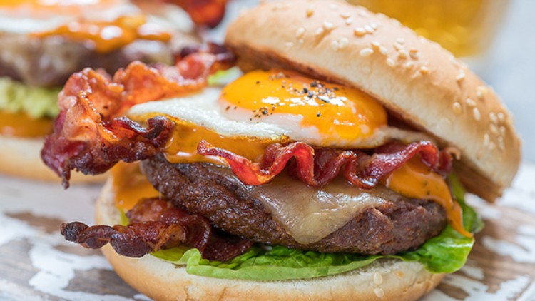 Burger mit Sesambrötchen, gebratenem Speck, Spiegelei, Rindfleischpatty, geschmolzenem Käse, grünen Salatblättern und Sauce.