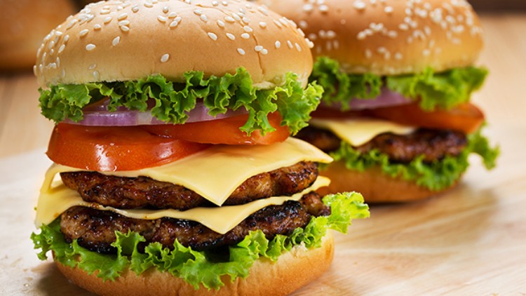 Zwei Hamburger mit Sesambrötchen, Salat, Tomate, Käse, Zwiebeln und zwei gegrillten Patties auf einem Holzuntergrund.