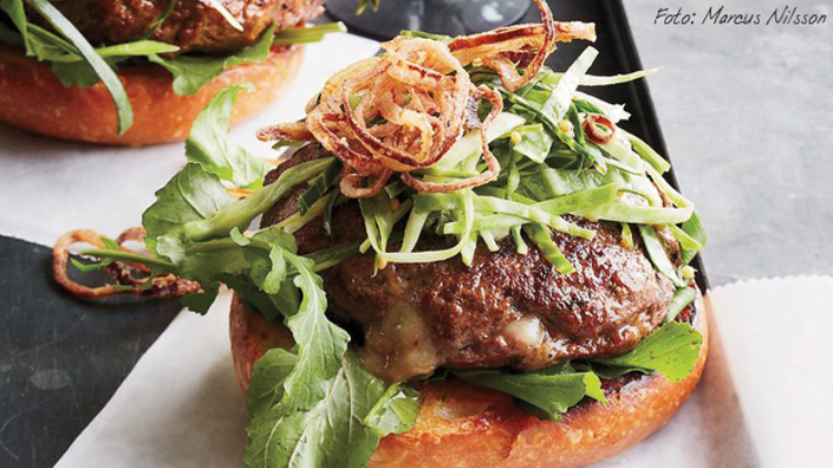 Burger mit Rucola, Krautsalat, knusprigen Zwiebelringen und gegrilltem Patty auf einem Brötchen-Unterteil auf Backpapier.