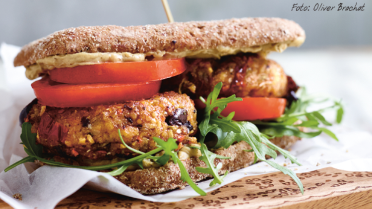 Vollkornbrot-Sandwich mit veganen Burger-Patties, Tomatenscheiben, Rucola und cremigem Aufstrich, angerichtet auf Backpapier.