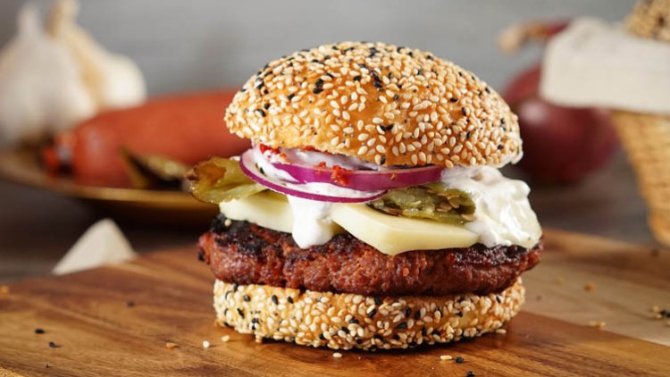 Burger mit Sesambrötchen, gegrilltem Patty, Käse, Zwiebeln, eingelegten Gurken und weißem Dressing auf Holzbrett.
