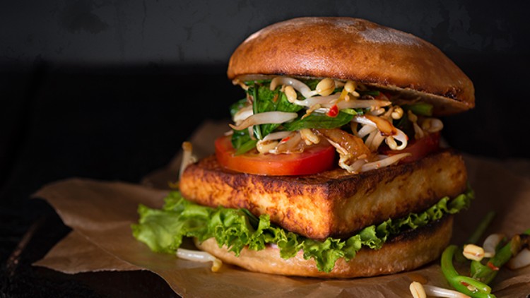Ein Burger mit gebratenem Tofu, Salat, Sprossen, Tomatenscheiben und weiteren frischen Zutaten auf braunem Papier.