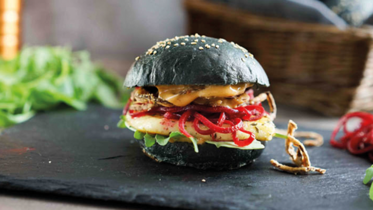 Ein Burger mit schwarzem Brötchen, Rucola, Halloumi, roten Zwiebeln, Soße und gebratenen Pilzen liegt auf einer Schieferplatte.