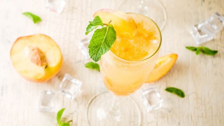 Ein Glas mit Pfirsich-Eisgetränk, dekoriert mit Minzblatt und Pfirsichscheibe, steht auf hellem Untergrund mit Eiswürfeln und Minze.