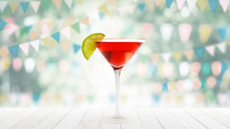 Rotes Getränk in einem Cocktailglas mit Limettenscheibe auf hellem Holztisch, bunte Wimpel im Hintergrund.