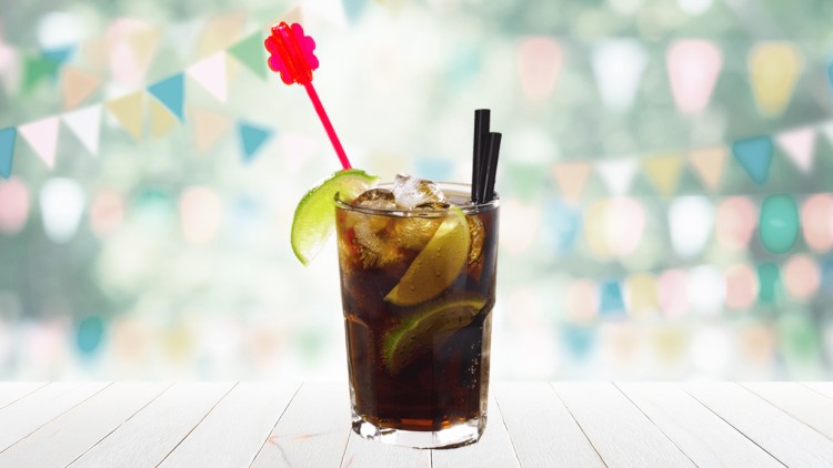 Ein Glas mit Cola, Eiswürfeln, Limettenscheiben, einem roten Rührstab und schwarzen Trinkhalmen auf weißem Holztisch.