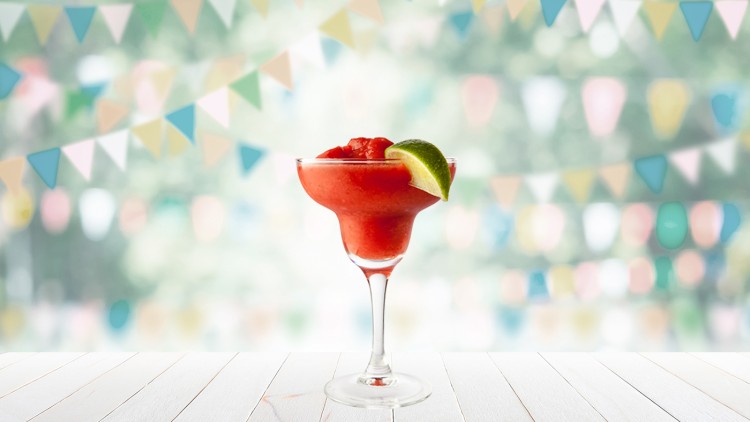 Rotes, cremiges Getränk in einem Kelchglas mit Limettenscheibe, im Hintergrund bunte Wimpelketten und verschwommene Natur.