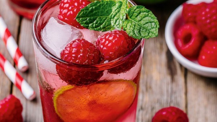 Ein Glas mit Himbeerlimonade, Eiswürfeln, Limettenscheiben und Minzblättern steht auf Holz, daneben Himbeeren und ein Limettenstück.