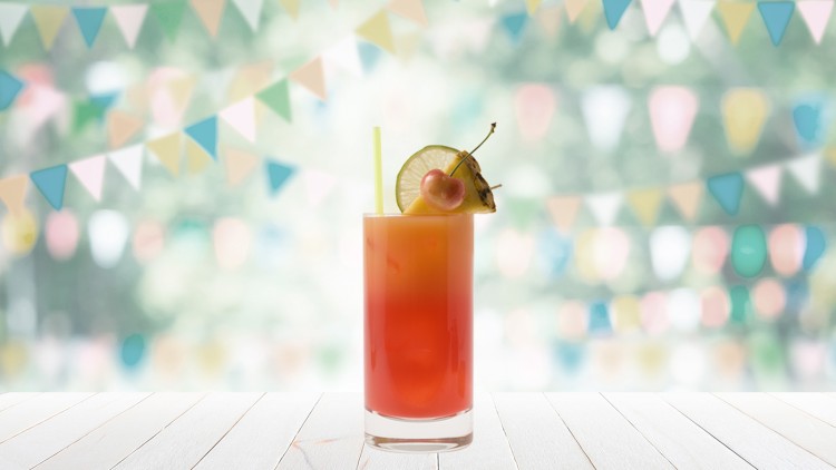 Ein Glas mit orangem Cocktail, Limettenscheibe, Kirsche und Ananas garniert, steht vor einer Wand mit bunten Wimpeln.