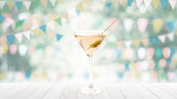 Ein Martini-Glas mit heller Flüssigkeit und drei Oliven auf einem Holzuntergrund, im Hintergrund bunte Wimpelketten unscharf.