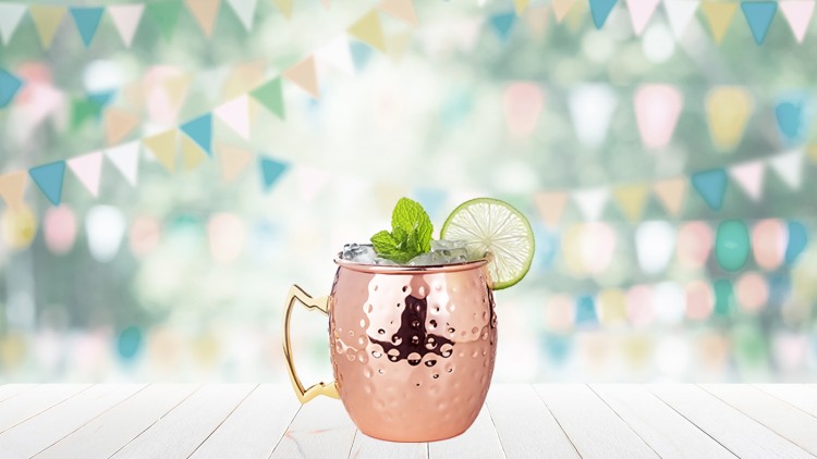 Kupferfarbener Becher mit Eiswürfeln, Minze und Limettenscheibe auf Holztisch, im Hintergrund bunte Wimpelgirlanden.
