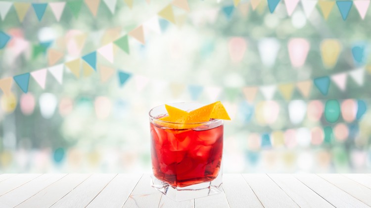 Ein Glas mit rotem Getränk, Eiswürfeln und Orangenscheiben steht auf einem hellen Holztisch, im Hintergrund bunte Wimpel.