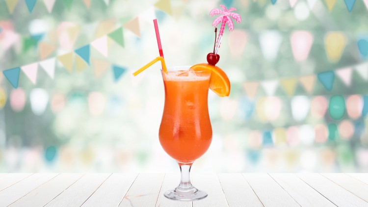 Ein orangener Cocktail mit Eis, Orangenstück, Kirsche, zwei Trinkhalmen und Deko-Palme steht auf weißem Holztisch.