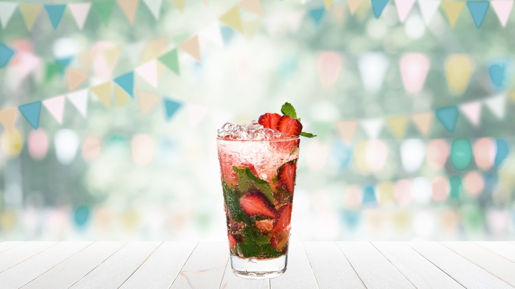 Ein Glas mit rosa Getränk, Erdbeeren, Minzblättern und Eiswürfeln, auf hellem Tisch, im Hintergrund bunte Wimpel.