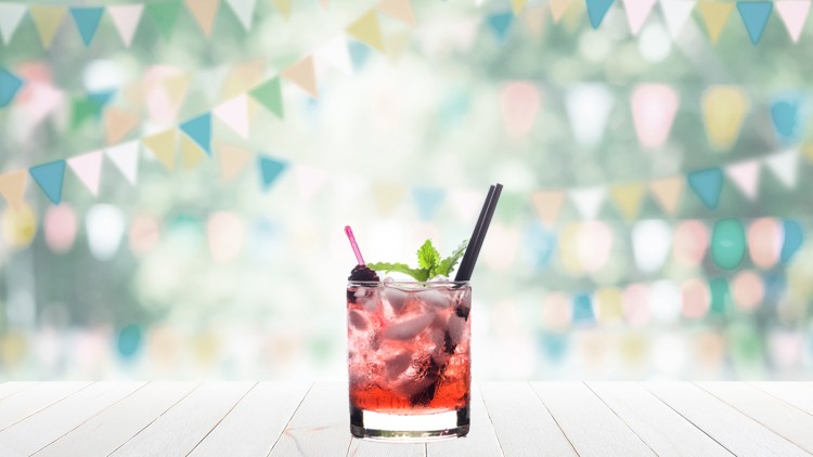 Ein Glas mit rotem Getränk, Eiswürfeln, Minzblättern, schwarzem Strohhalm und rosa Rührstäbchen vor unscharfer Wimpelkette.