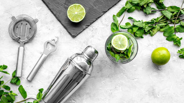 Cocktailshaker, Barsieb, Flaschenöffner, Limetten, Minze und ein Glas mit Limettenscheibe auf grauem Hintergrund.