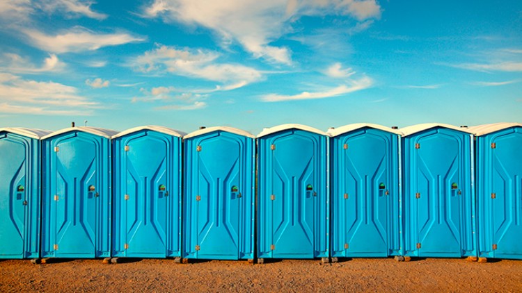 Eine Reihe blauer mobile Toilettenkabinen steht auf einem sandigen Untergrund vor einem Himmel mit weißen Wolken.