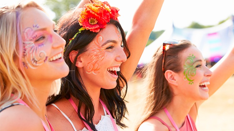 Drei Personen in sommerlicher Kleidung heben die Arme, eine trägt einen Blumenkranz, im Hintergrund ist eine Wiese erkennbar.