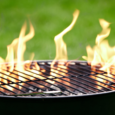 Ein Grillrost mit offenen Flammen, im Hintergrund unscharf grüne Wiese.