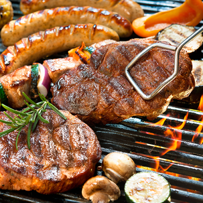 Mehrere gegrillte Fleischstücke, Würstchen und Gemüse wie Zucchini, Paprika und Champignons liegen auf einem heißen Grillrost.