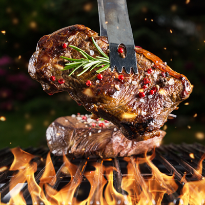 Ein saftiges Steak mit Gewürzen und Rosmarinzweig wird mit einer Grillzange über einem Grill mit offenen Flammen gehalten.