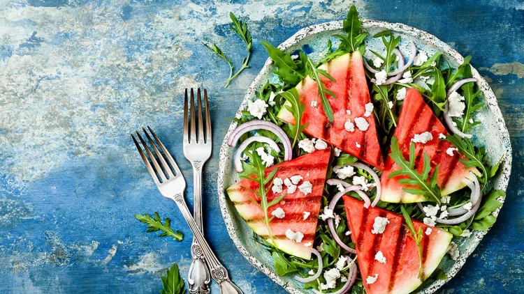 Ein Teller mit gegrillten Wassermelonenstücken, Rucola, roten Zwiebelringen und zerbröckeltem weißen Käse, daneben zwei Gabeln.