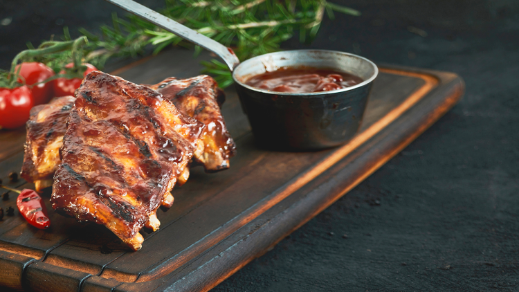 Gegrillte Rippchen mit glänzender Marinade auf einem Holzbrett, daneben eine Schale mit Sauce, Chili und frische Tomaten.