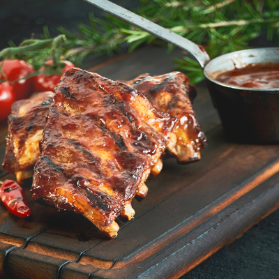 Gegrillte Rippchen mit glänzender Marinade auf einem Holzbrett, daneben eine Schale mit Sauce, Chili und frische Tomaten.