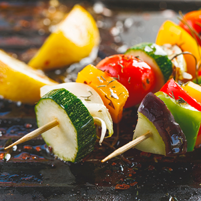 Gemüsespieße mit Zucchini, Tomaten, Paprika und Auberginen liegen auf einem Grillrost, daneben Stücke einer Zitrone.