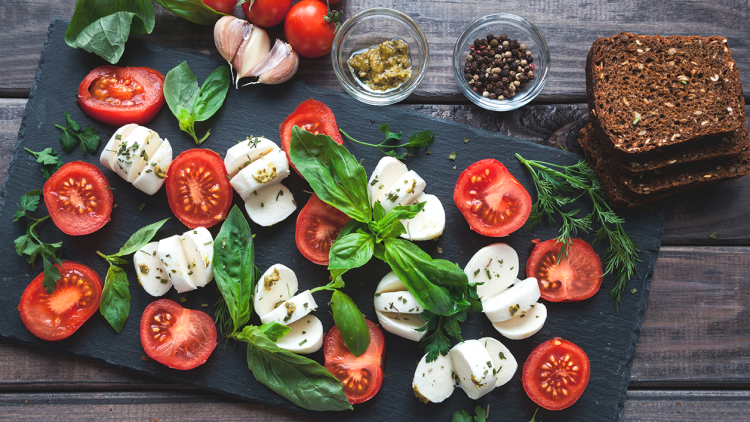 Tomatenscheiben, Mozzarella, frisches Basilikum und Kräuter auf einer dunklen Platte, daneben Gewürze, Knoblauch und Brot.