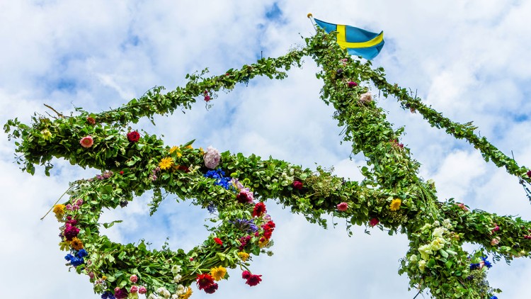 Mit Blumen und Blättern geschmückte Stange in Kreuzform mit schwedischer Flagge an der Spitze vor einem bewölkten Himmel.