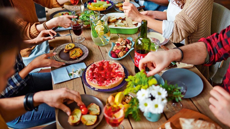 Menschen sitzen an einem gedeckten Holztisch mit Speisen wie Kuchen, Salat, Nüssen, Kräutern und Getränken.
