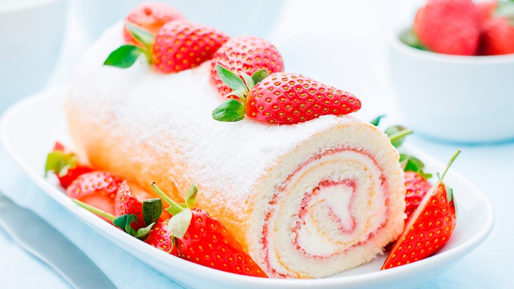 Biskuitrolle mit cremiger Füllung, Erdbeerstreifen und ganzen Erdbeeren, dekoriert mit Puderzucker, auf weißem Teller.