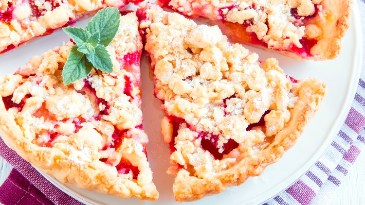 Vier Stücke Streuselkuchen mit roter Füllung und einem Minzblatt auf weißem Teller, darunter eine lilafarben gestreifte Serviette.