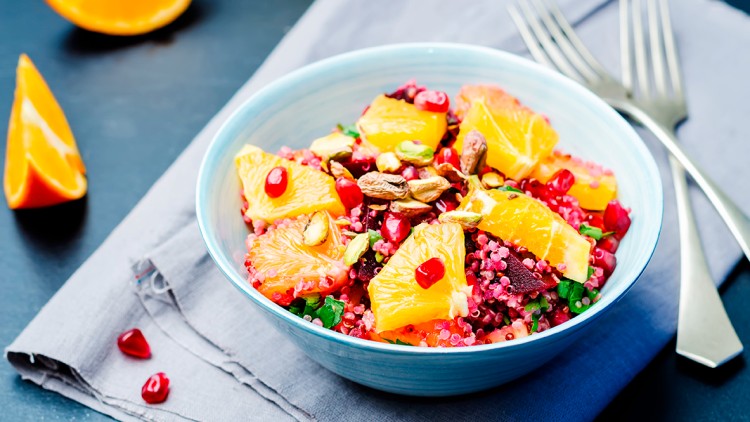 Schüssel mit Quinoa, Orangenscheiben, Granatapfelkernen, Pistazien und Kräutern auf einem Stofftuch, daneben zwei Gabeln.