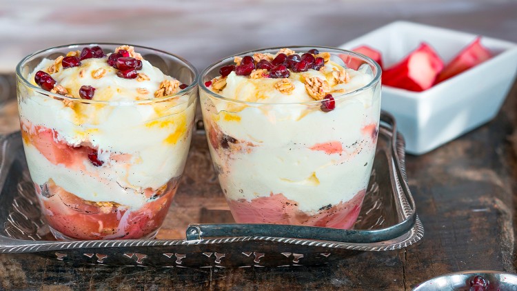 Zwei Dessertgläser mit Schichten aus Creme, Rhabarberkompott, Granatapfelkernen und Müsli auf altem Holzuntergrund.